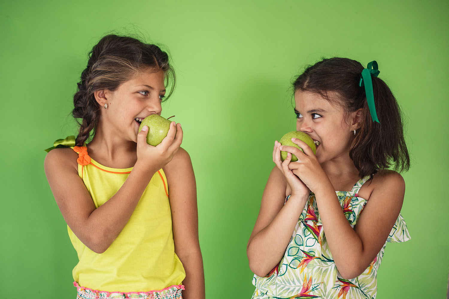 Front view of two girls with ponytails looking at each other and eating green organic apples.